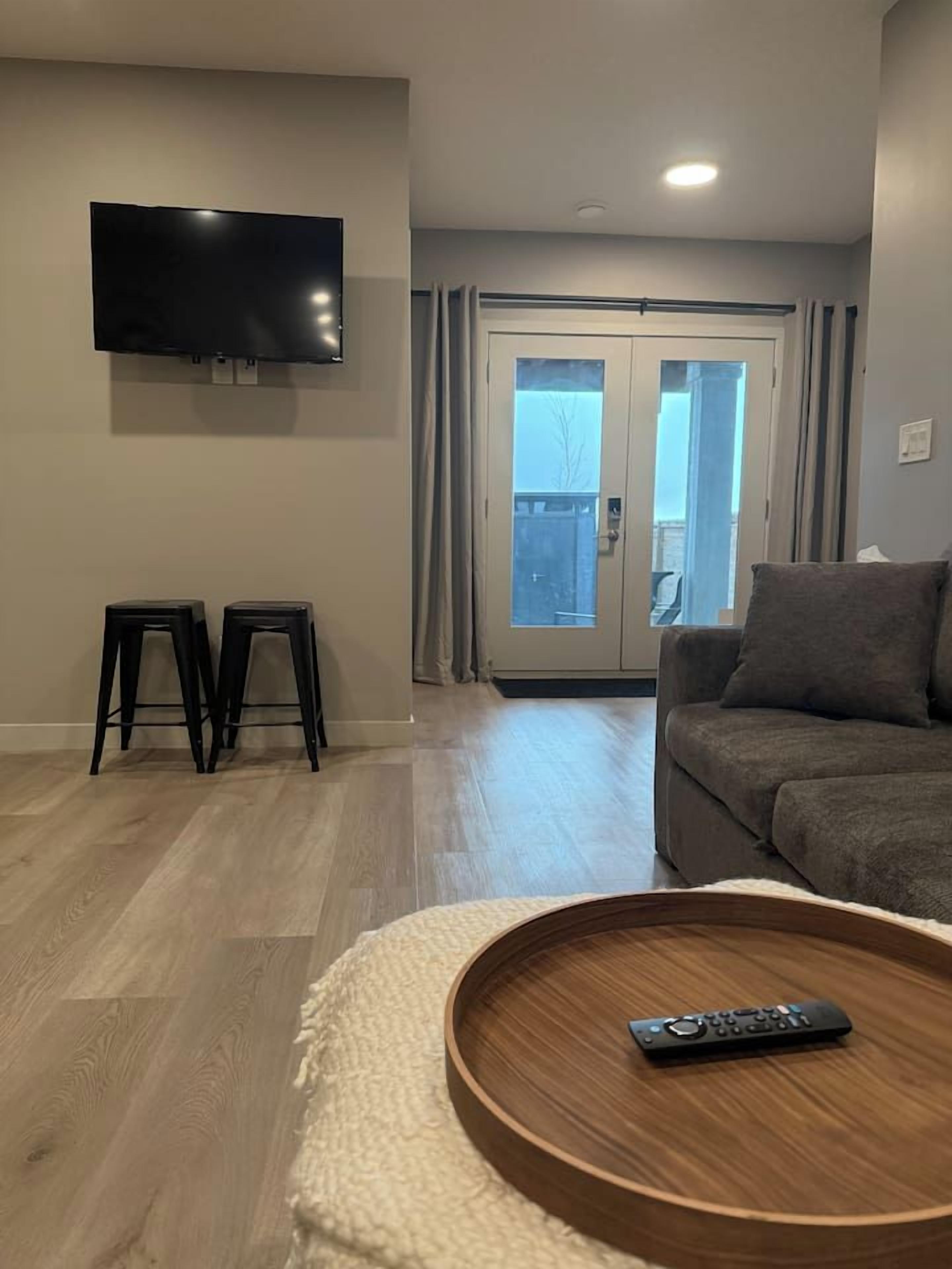 Ein Wohnzimmer mit einem an der Wand befestigten Flachbildfernseher, einem grauen Sofa, einem runden Couchtisch mit einer Fernbedienung, zwei schwarzen Hocker und einer Schiebetür zu einem Außenbereich.