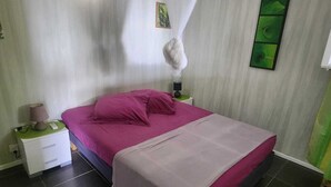 A bedroom with a bed, a nightstand, a lamp, a wall-mounted picture, and a curtain. - T2 CREOLE apartment by the pool (Sainte-Luce)