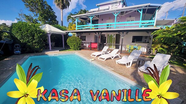 A swimming pool with lounge chairs, a covered patio, and a house with a blue balcony. - T2 CREOLE apartment by the pool (Sainte-Luce)
