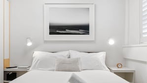 A neatly made bed with white linens, a framed picture of the ocean on the wall, a bedside lamp, and a small bedside table with a clock. - Bay View in Rushcutters Bay (Sydney)