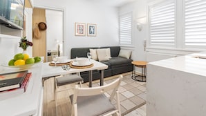 A modern living room with a grey sofa, a dining table with chairs, a TV mounted on the wall, and a large window with blinds. - Bay View in Rushcutters Bay (Sydney)