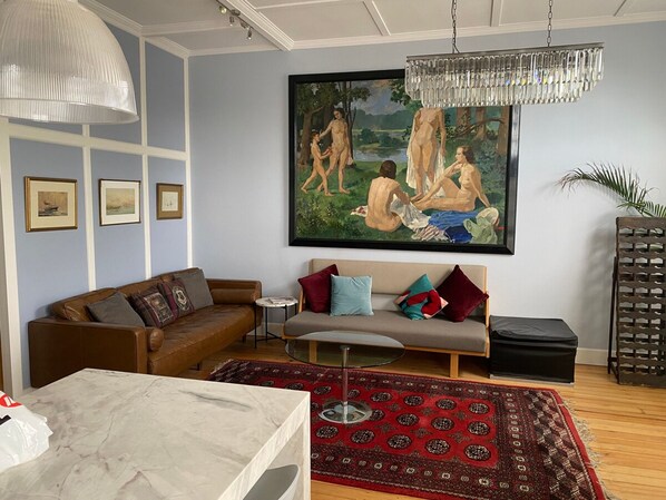 A living room with a brown leather sofa, a grey sofa with colorful cushions, a red patterned rug, and a large painting of nude figures on a wall. - Duffotel Paddington Exec 2-bedroom Apartment (Sydney)