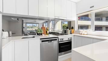 A modern kitchen with white cabinets, a dishwasher, an oven, and a window with a city view.