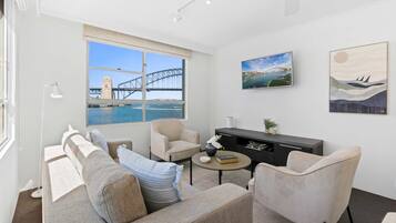A modern living room with a sofa, armchairs, a coffee table, and artwork on the walls. A large window offers a view of a bridge and water.