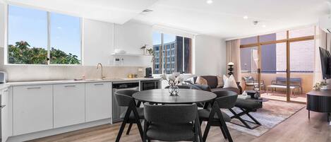 A modern kitchen with white cabinets, a dining area with a round table and chairs, and a living area with a sofa and a TV.