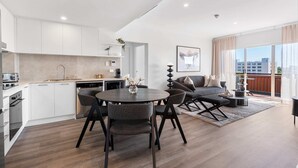 A modern apartment with a kitchen, dining area, and living room. It features a large window with a city view, a dining table with chairs, and a sofa set. - Camperdown Cozy Corner 10 (Sydney)