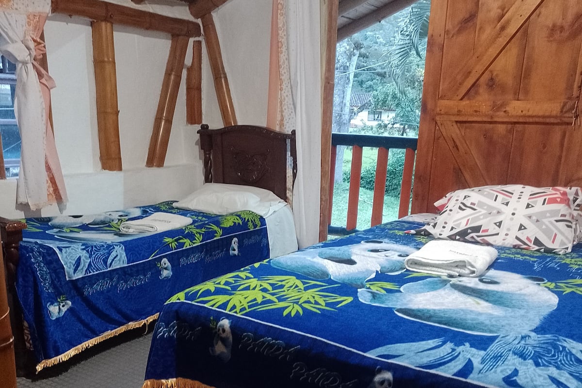 Quarto com duas camas, cada uma com colcha azul estampada com pandas, vigas de madeira no teto e vista para o verde lá fora.