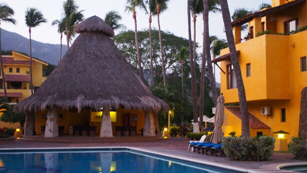 A pool area with a thatched-roof structure, palm trees, and a yellow building with balconies. - Apartment in Club Santiago (Manzanillo)