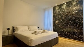 Une chambre d’hôtel moderne avec un grand lit, du linge de lit blanc, un mur décoratif en marbre et une table de chevet avec une lampe.