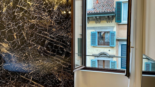 Una habitación con pared de mármol y ventana que da a un edificio con contraventanas azules.