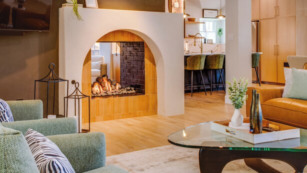 A modern living room with a fireplace, green upholstered sofas, a glass coffee table, and a kitchen area in the background. - Family & Dog-Friendly Luxury Tudor Home in Richardson, TX w/ Pool and Spa (Richardson)