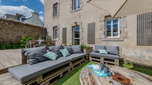 A courtyard with a wooden deck, two grey sofas with cushions, a rustic coffee table, and a stone wall. - Holiday Home 'Maison 8 Pers Plein Centre Ville' with Private Terrace, Garden & Wi-Fi (Lesneven)