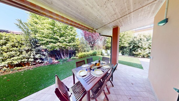 An outdoor patio with a table set for dining, surrounded by chairs, and a view of a well-maintained garden.