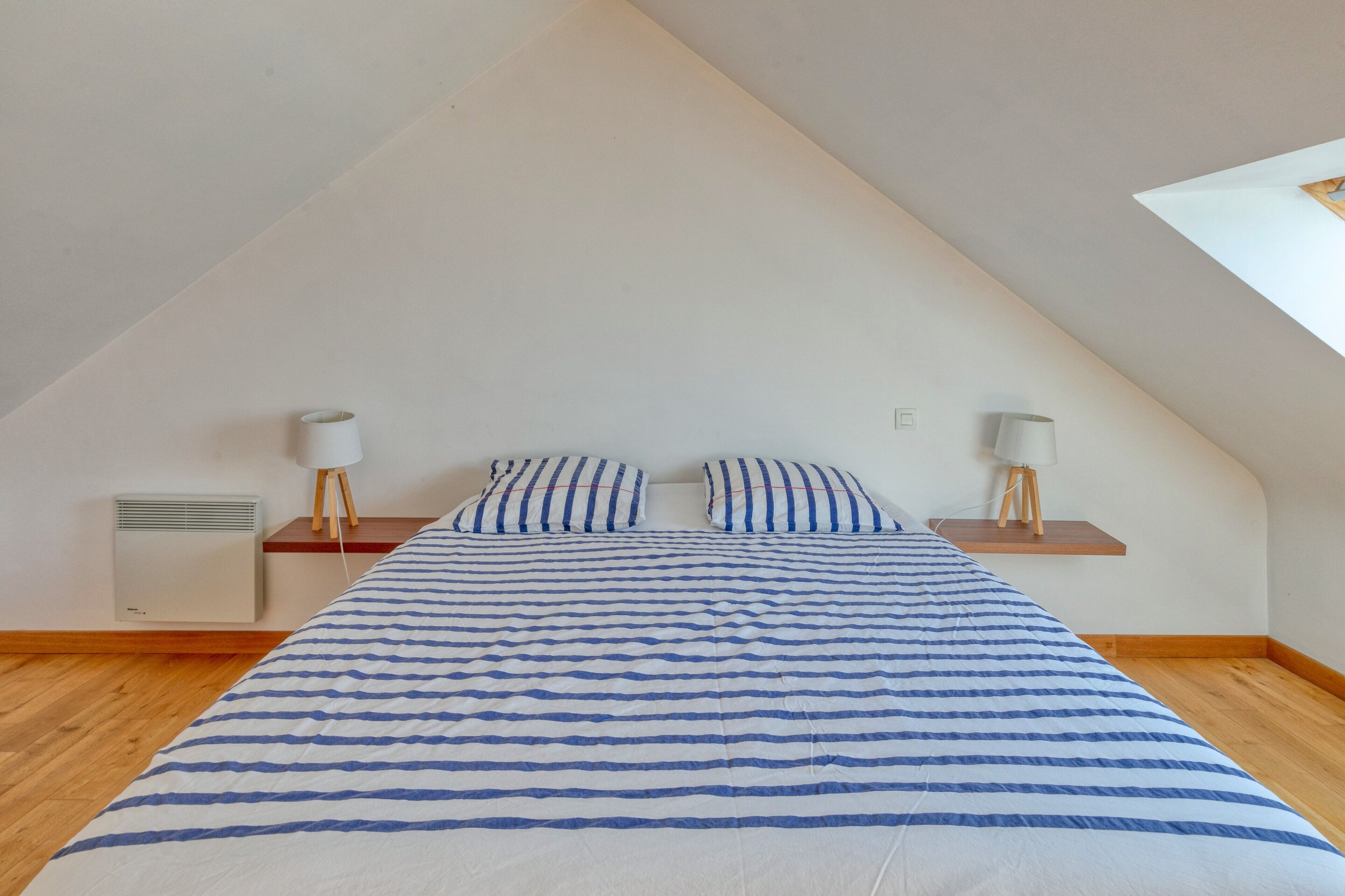 Une chambre avec un plafond en pente, un lit recouvert d’une couvre-lit à rayures, deux lampes de chevet et un radiateur fixé au mur.