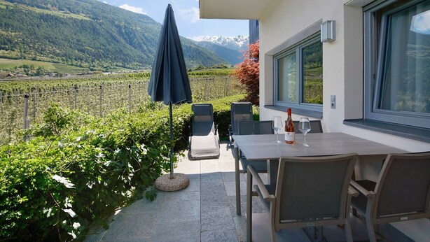 Un patio con una mesa y sillas, una botella de vino y vistas a un viñedo y montañas.