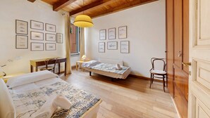 A room with a wooden ceiling, a bed with a floral bedspread, a desk with a chair, a small table with a lamp, and a chair.