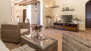 A living room with wicker furniture, a glass coffee table, a TV on a wooden stand, and a hallway leading to another room. - Holiday Home 'Pescadores' with Sea View, Private Terrace and Air Conditioning (Son Moll)