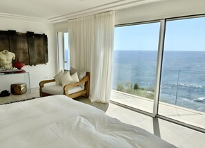 Un dormitorio con una cama grande, una silla de madera, una mesita y vistas al océano.