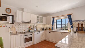 A kitchen with white cabinets, a microwave, a coffee maker, and a window with blue curtains. - Holiday Home 'Leo' with Sea View, Wi-Fi and Air Conditioning (Faro)