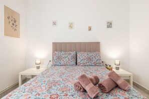 Una cama bien hecha con ropa de cama floral, dos mesitas de noche con lámparas, y una pared con cuatro pequeñas obras de arte.