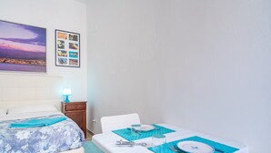 Habitación de hotel con una cama, una mesa para dos personas, una mesita de noche con lámpara, y una pared con un cuadro y un cartel.