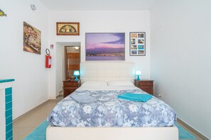Une chambre à coucher comprenant un lit, deux tables de chevet, une commode, un extincteur, un tableau encadré et une porte donnant sur une autre pièce.