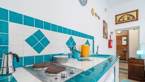 Una cocina con estufa, fregadero y azulejos azules. Hay platos apilados en la encimera, una tetera y un recipiente amarillo.