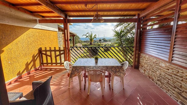 Eine überdachte Terrasse mit Tisch und Stühlen, Blick ins Grüne und eine Steinmauer.