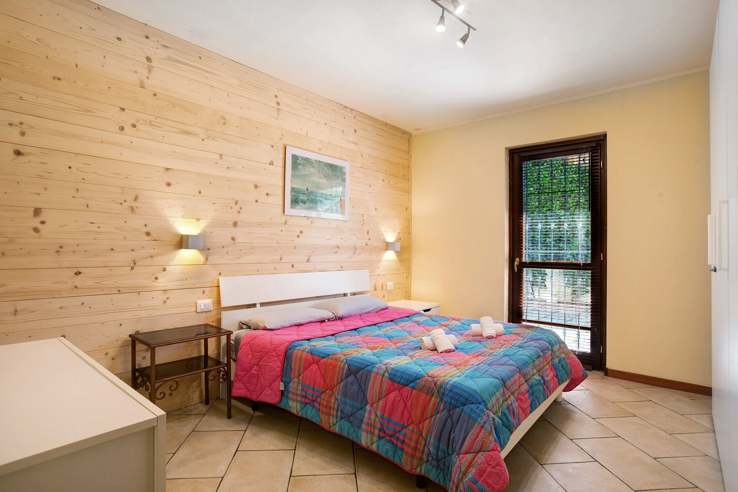 A bedroom with a wooden wall, a bed with a colorful quilt, two bedside lamps, and a window with blinds.