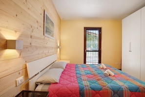 A bedroom with a wooden headboard, a bed with a colorful quilt, a window with blinds, and a white wardrobe.