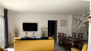 A living room with a yellow sofa, a flat-screen TV, a dining area with a table and chairs, and a decorative wall.