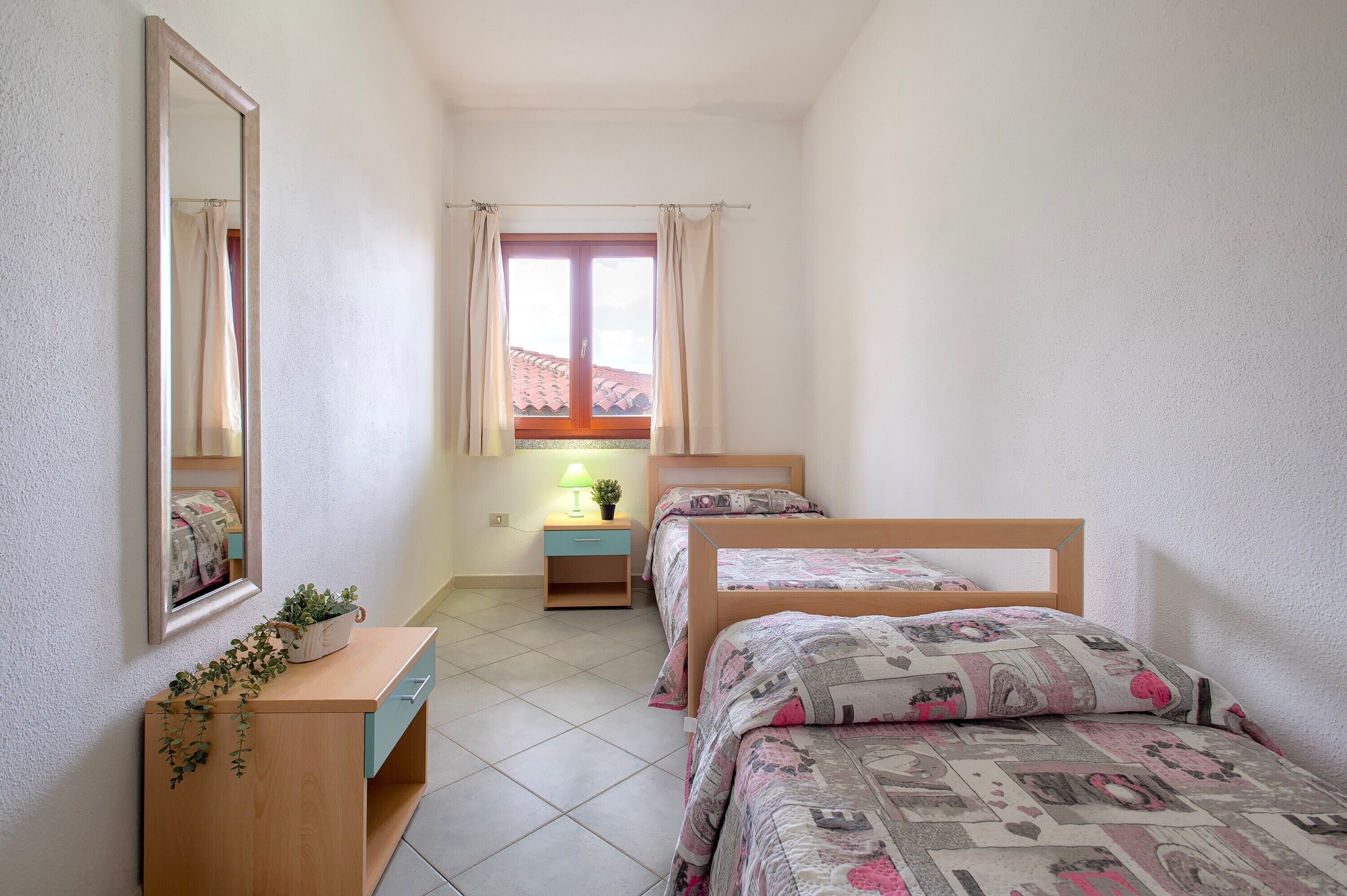 Une chambre avec deux lits, une table de chevet avec une lampe, un miroir et une fenêtre avec des rideaux.