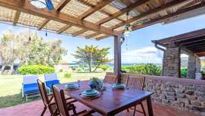 Un patio al aire libre con una mesa de madera lista para comer, rodeado de sillas, un muro de piedra y vista al océano.