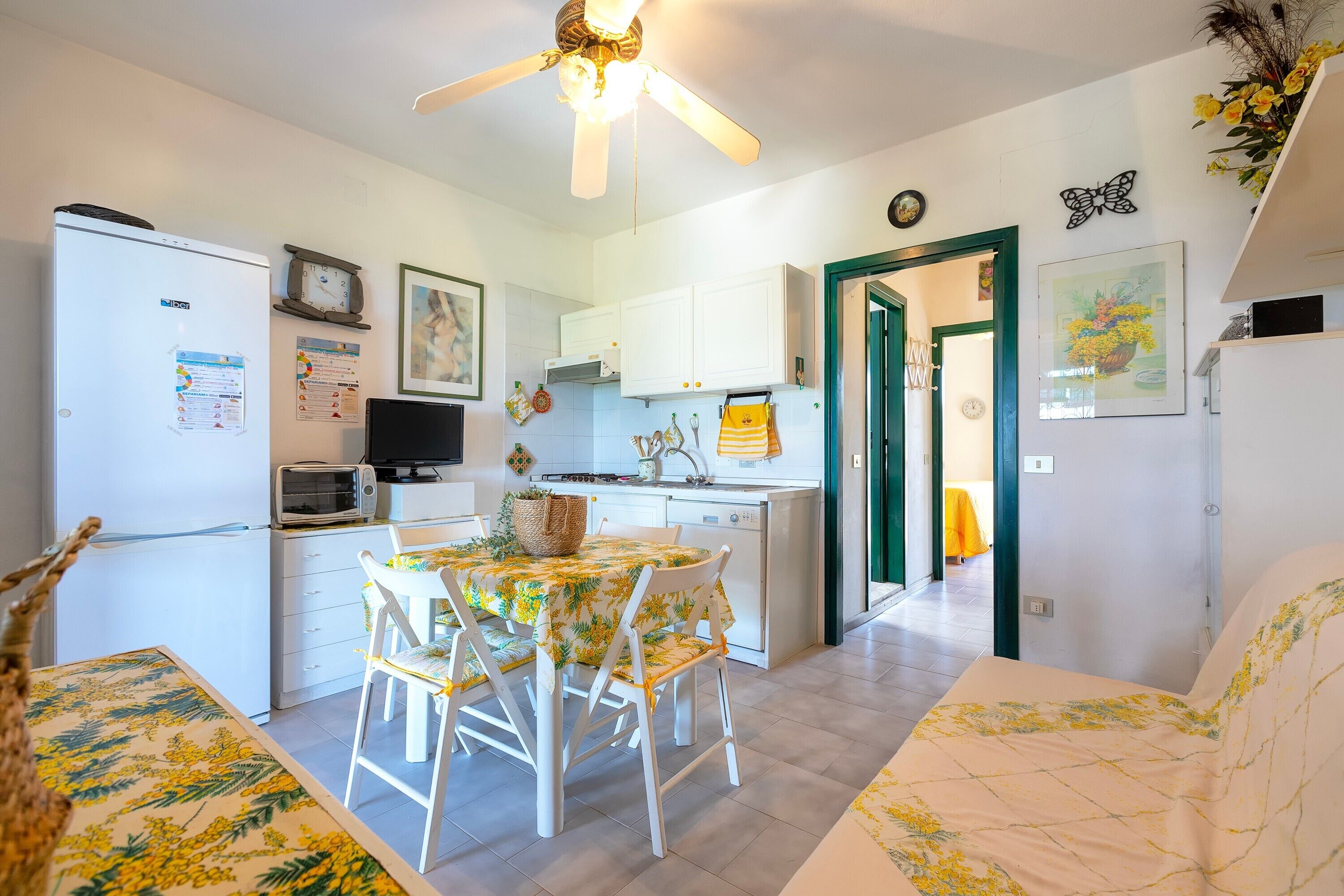 A compact kitchen with white cabinets, a dining table with chairs, a ceiling fan, and a small TV.