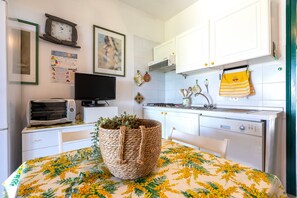 Une cuisine avec des meubles blancs, un grille-pain, un évier et une table recouverte d’une nappe à motifs floraux.