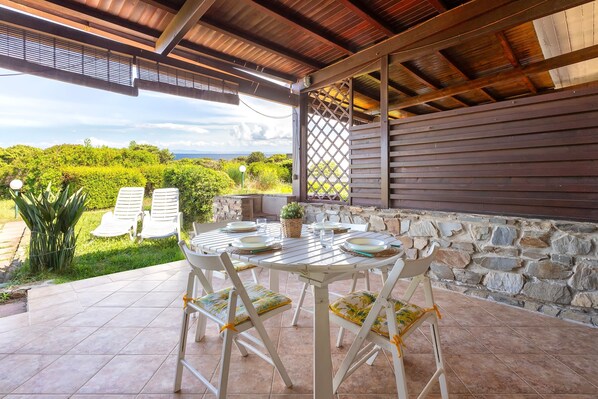 An outdoor patio with a table set for dining, surrounded by chairs, a stone wall, and a view of greenery.