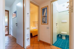 Quarto de hotel com cama, banheiro com piso azul e um corredor que dá para outro quarto.
