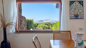 Una habitación con vistas a montañas y al mar, una mesa con sillas, un florero con plantas secas y un azulejo decorativo en la pared.