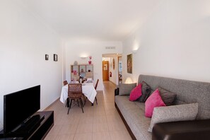 A living room with a flat-screen TV, a grey sofa with pink cushions, a dining area with a white table, and a hallway leading to another room.