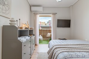 A bedroom with a bed, a dresser, a TV, and a view of a balcony with a chair and a small garden.