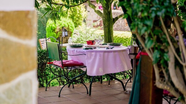An outdoor dining area with a table set for two, surrounded by greenery and a stone wall.
