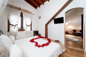Una camera da letto con un letto, tende bianche, una televisione e vista su un soggiorno attraverso un arco.