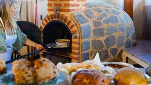 A brick oven with a stone exterior, a person using a tool, and various baked goods in the foreground. - Room 'Doble Confort Dalia' with Mountain View, Shared Terrace and Wi-Fi (Rubayo)