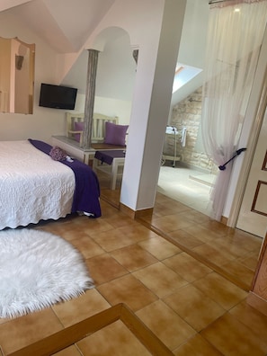 A bedroom with a large bed, a small table, a chair, a TV, and a bathroom with a shower. - Double Violeta Room with Mountain Views, Shared Terrace, and Wi-Fi (Rubayo)