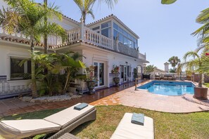 Una casa de dos pisos con piscina, mobiliario exterior y plantas tropicales.