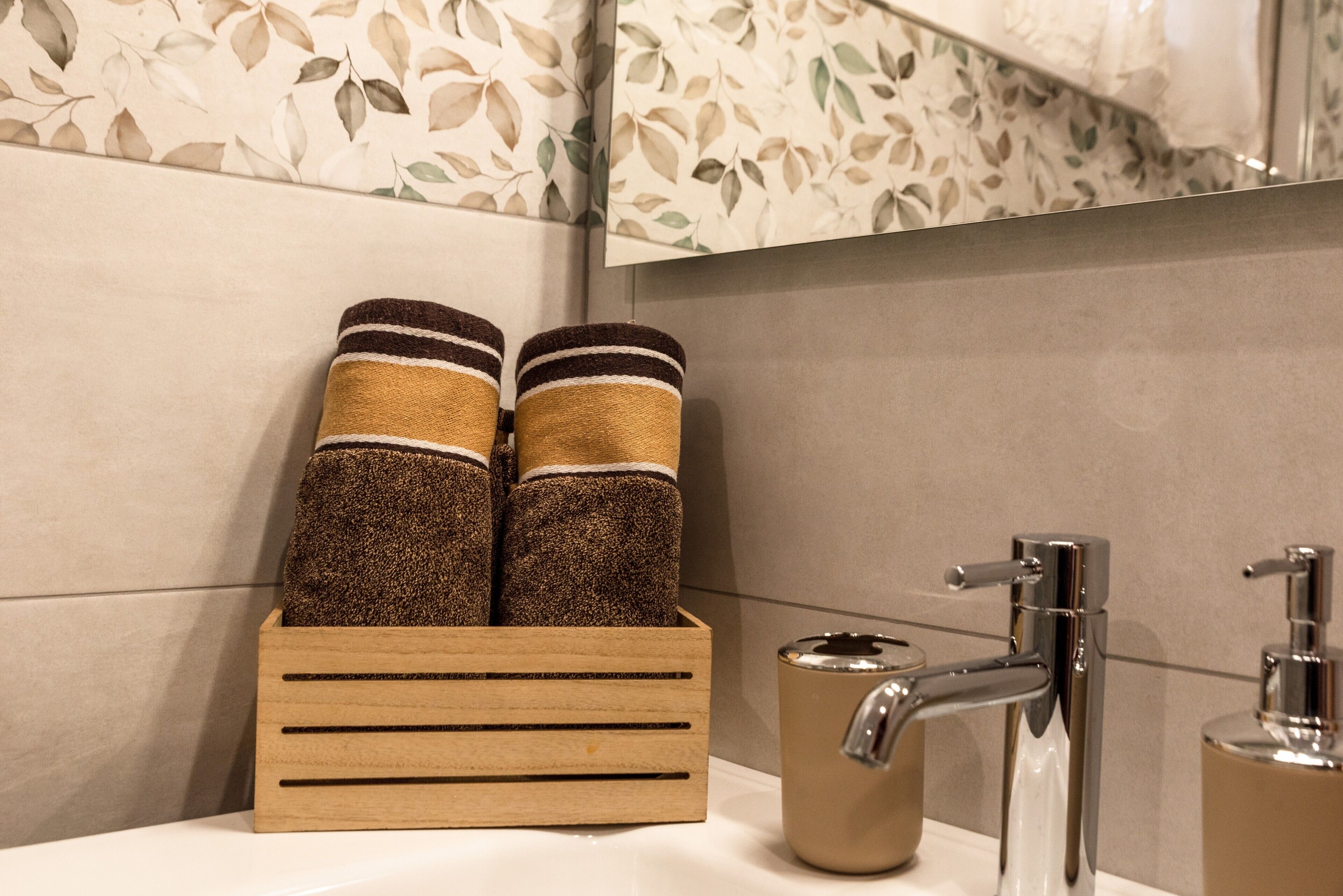 A wooden towel rack with two towels, a bathroom sink with a chrome faucet, and a mirror with a leaf-patterned backsplash.