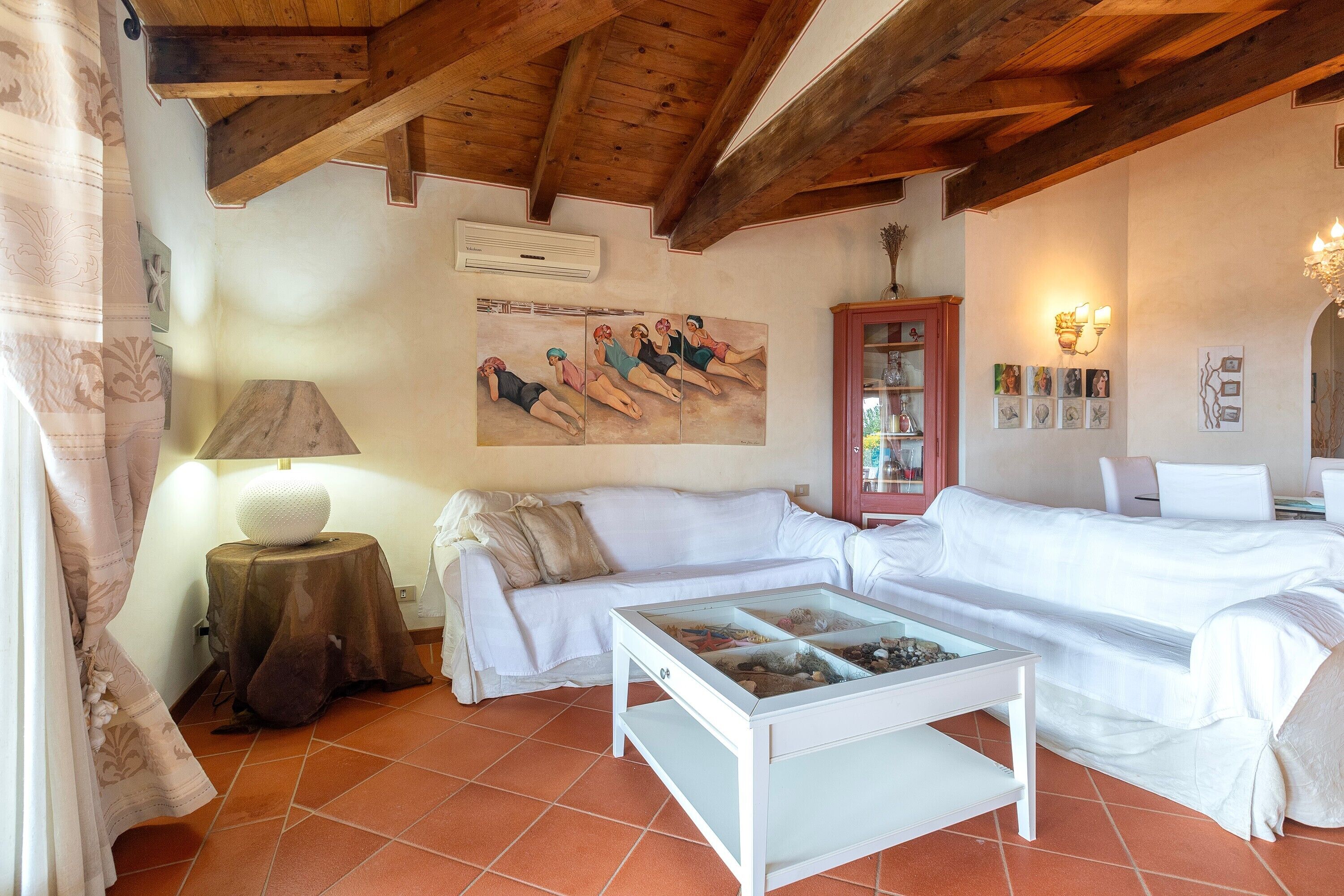 A room with a white sofa, a glass-top coffee table, a wooden ceiling, and a wall-mounted air conditioner.