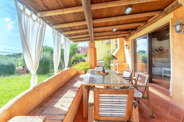A covered patio with a wooden table and chairs, a glass door leading to an outdoor area, and a view of greenery.