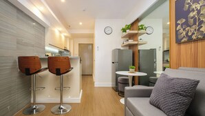 A modern living room with a grey sofa, two wooden bar stools, a small round table with a plant, and a wall-mounted clock. - Great Holiday Vacation: 1BR Condo w/ 2 Queen Beds (Quezon City)
