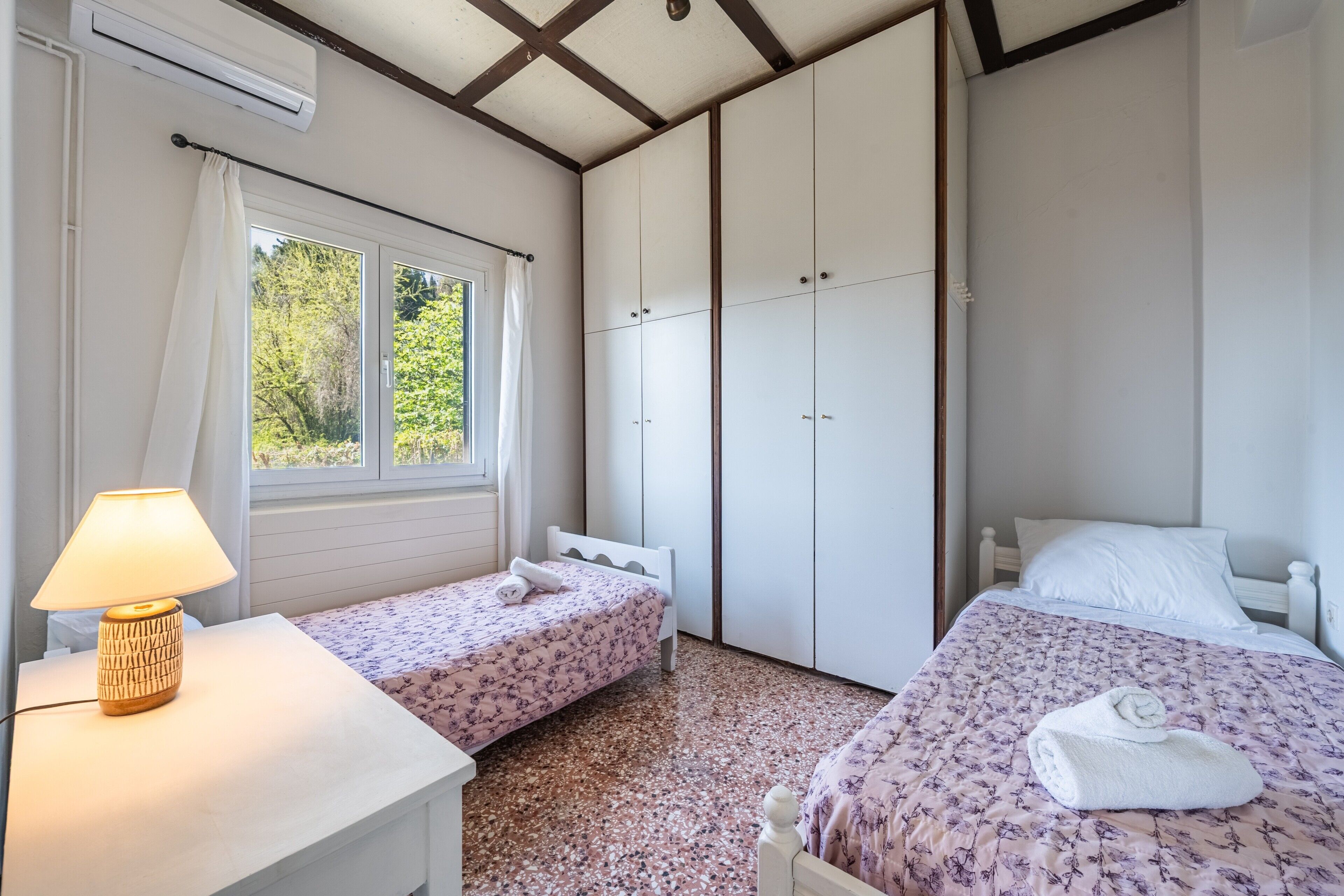 A bedroom with two beds, a window with curtains, an air conditioner, and a bedside lamp.